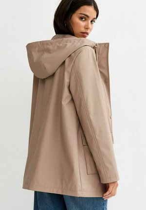 Vrouw die zich omdraait, gekleed in een beige regenjas met capuchon en drukknopen, en een blauwe spijkerbroek, tegen een effen witte achtergrond.