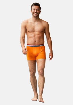 Lachende shirtloze man die op blote voeten loopt en feloranje boxershorts draagt met een "HAPPY SHORTS" tailleband op een witte achtergrond.