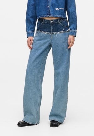 Szerokie dżinsy w dwóch odcieniach niebieskiego, z ciemnym dżinsem u góry i jasnym u dołu, ozdobione srebrnymi ćwiekami wzdłuż linii talii, noszone z czarnymi butami.