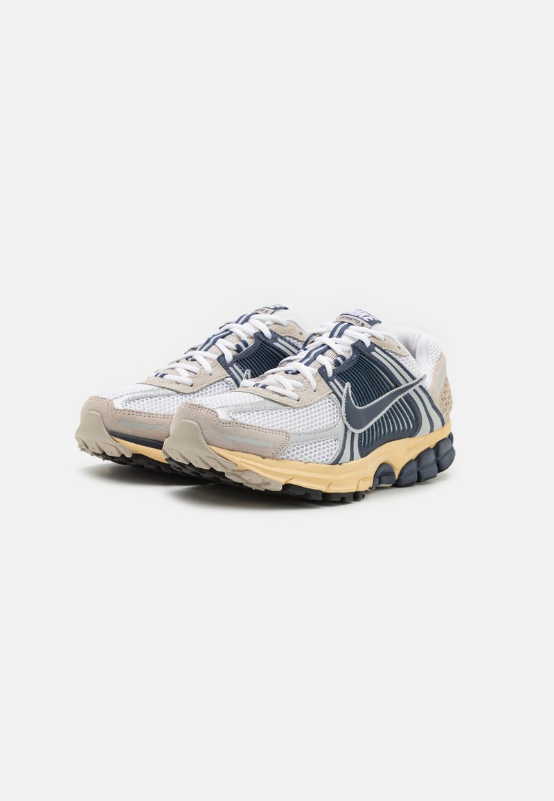 Zapatillas con una parte superior de malla y textil en blanco y azul marino, con acentos en beige y una suela de goma texturizada para tracción.