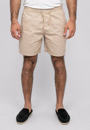 Homme portant un short beige à cordon, une chemise blanche et des chaussures noires sans lacets, debout devant un fond uni.