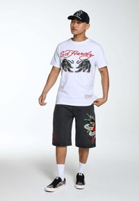 T-shirt bianco con stampa di pantera nera, pantaloni corti in denim neri con design ispirato ai tatuaggi colorati, cappellino nero e sneaker con motivo.