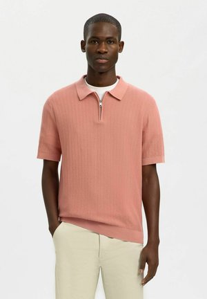 Man wearing salmon pink short-sleeve knit polo with zipper and beige pants, standing with one hand in pocket against plain background.