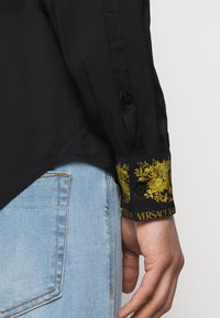 Camisa negra con un puño estampado que presenta diseños florales dorados y botones. El puño contrasta con el cuerpo negro sólido, mostrando textura y detalle.