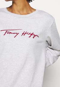 Šedá mikina s kulatým výstřihem. Na přední straně má červeně vyšívané logo „Tommy Hilfiger“. Měkká, texturovaná látka.