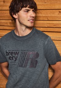 Grå, kortärmad t-shirt i mjukt tyg, med ett tryckt motiv i mörkare grått som visar texten "brewery" och stiliserat "R."