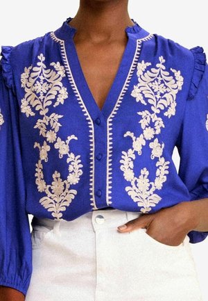 Sininen pusero, jossa valkoinen kukkaommel, V-kaula-aukko ja napit, röyhelöidyt olkapäät, valkoisten korkeavyötäröisten housujen kanssa, käsi taskussa.