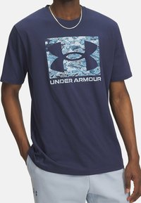 Camiseta de algodón azul marino con un logo de camuflaje azul claro de Under Armour en un cuadrado, cuello redondo y mangas cortas. Textura suave.