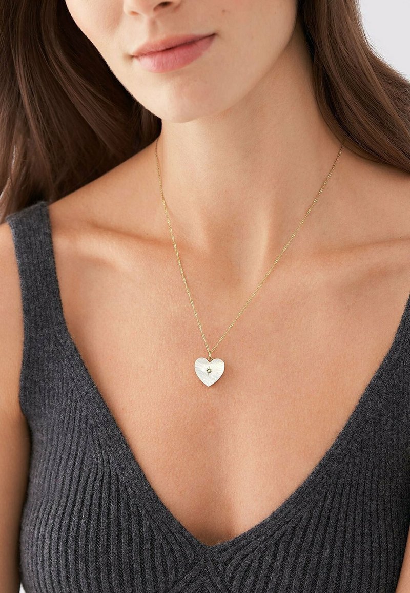 Collier chaîne en or avec un pendentif en forme de cœur texturé orné d'une petite pierre au centre, porté sur un haut côtelé gris foncé.