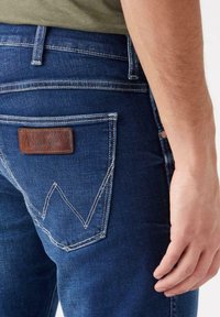 Los jeans de mezclilla de Wrangler en oscuro cuentan con una etiqueta de cuero en el bolsillo trasero con costuras blancas en un patrón de zigzag. Primer plano de la vista posterior.