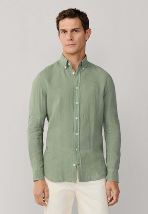 Hombre vestido con una camisa de manga larga abotonada de color verde claro y pantalones blanco hueso, de pie contra un fondo liso de color gris claro.