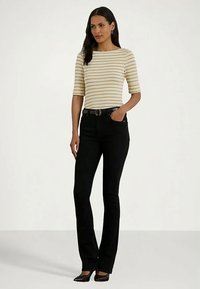 Frau steht da und trägt ein beige-weiß gestreiftes Oberteil mit Ellenbogenärmeln, einen schwarzen Gürtel, schwarze, figurbetonte Hosen und schwarze spitze Schuhe.