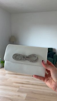 Λευκή δερμάτινη τσάντα clutch με λείο υφάσμα, διαθέτει μια μεγάλη διακοσμητική φιόγκο με πολύτιμους λίθους σε ασημί απόχρωση. Σχεδιασμένη με κλείσιμο τύπου καπάκι και ορθογώνιο σχήμα.