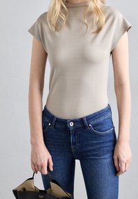 Beige top met korte mouwen en een hoge halslijn, gecombineerd met donkerblauwe slim-fit jeans. Het model houdt een zwarte handtas vast met een crèmekleurige binnenkant.
