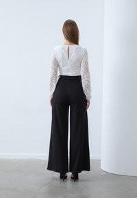 White lace long-sleeve top with a keyhole back, paired with wide-leg black trousers. Model wears black heels. Smooth fabric and structured fit.