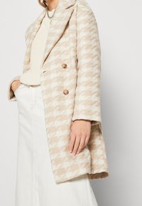 Beige kappa med hundtandsmönster av texturerad tyg; har en dubbelknäppt front och bruna knappar.