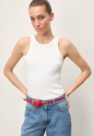Giovane donna con top bianco senza maniche e jeans blu, che indossa una cintura rossa con fibbia decorativa a forma di rosa, in posa con le mani sui fianchi.