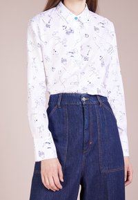 Camicia bianca con motivi astratti blu, chiusura a bottoni e un bottone di accento turchese. Abbinata a pantaloni in denim a vita alta.