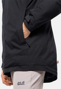 Schwarze wasserdichte Jacke mit hohem Kragen, verstellbaren Bündchen und Taschen. Begleitet von hellgrauen Hosen mit einem Markenlogo.