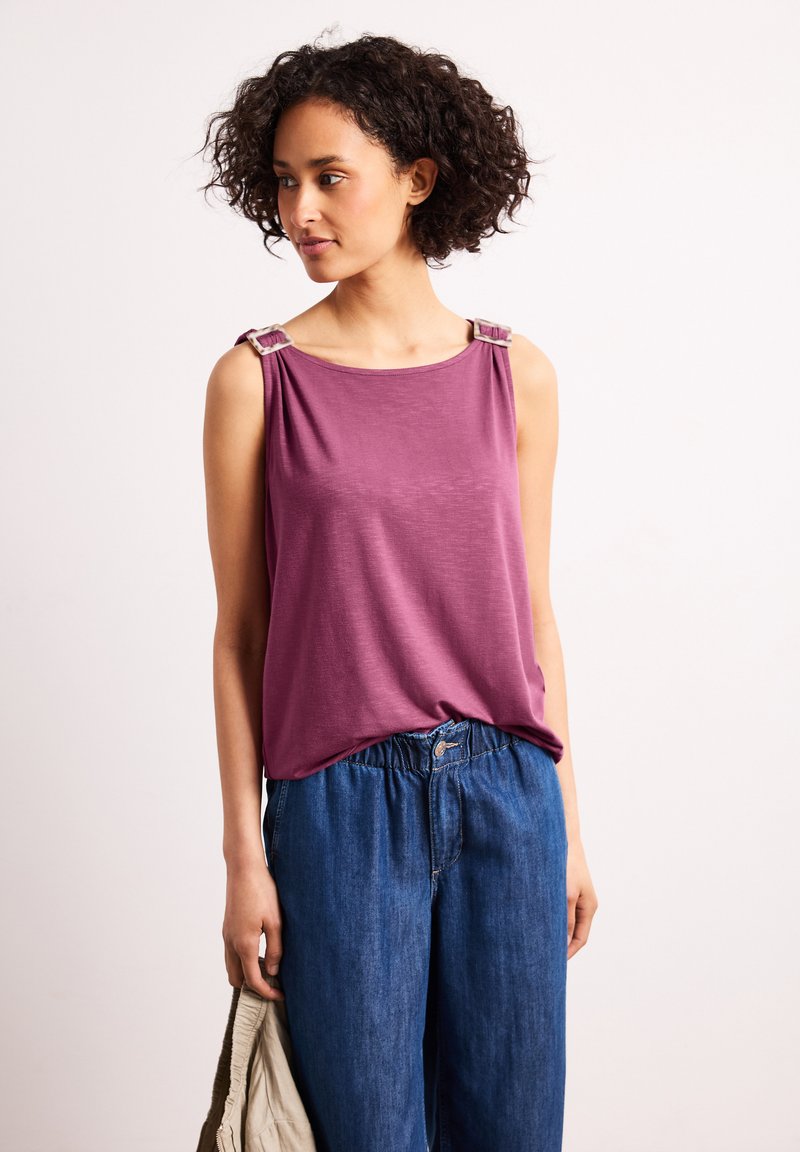 Femme aux cheveux bouclés portant un haut violet sans manches avec des bretelles à boucles et un jean bleu ample, tenant une veste beige dans la main gauche.
