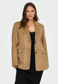 Beiger Blazer aus Kunstwildleder mit Reverskragen, Vordertaschen mit Klappen und Zweiknopfverschluss, kombiniert mit einem schwarzen, taillierten Oberteil und Rock.