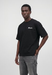 Homme noir portant un t-shirt noir avec le texte « Dropsize » sur la poitrine et un jean gris délavé, debout devant un fond gris uni.