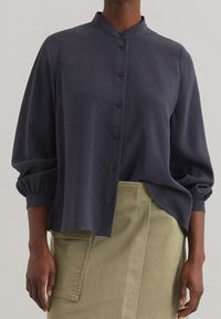 Dunkelgraues, kragenloses Button-up-Shirt mit lockeren Ärmeln und lässiger Passform, aus weichem Stoff. Kombiniert mit einem olivgrünen Rock mit Taschen.