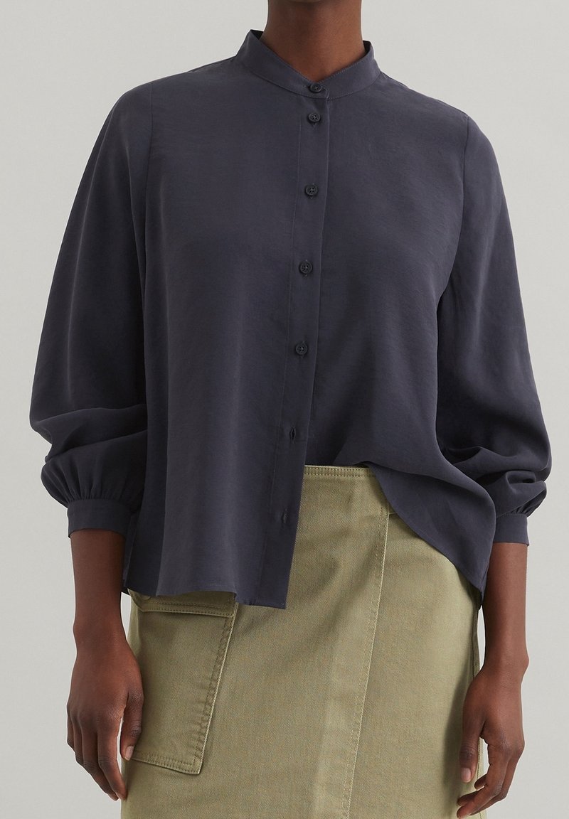 Dunkelgraues, kragenloses Button-up-Shirt mit lockeren Ärmeln und lässiger Passform, aus weichem Stoff. Kombiniert mit einem olivgrünen Rock mit Taschen.