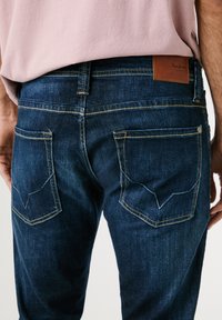 Mörkblå denimjeans med kontrasterande gul söm, med snedställda bakfickor och en brun läderetikett vid midjan.