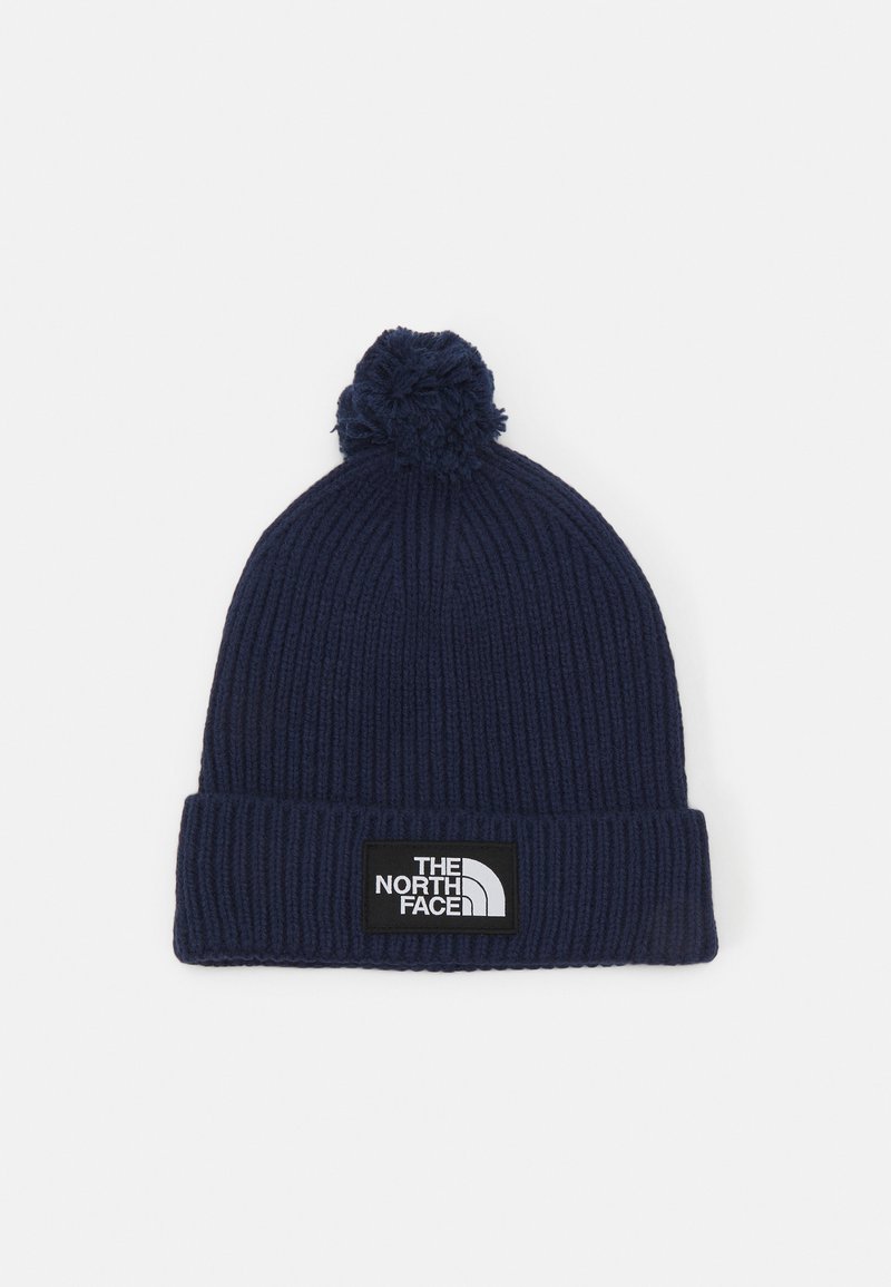 Marineblå beanie-hat med ribbet tekstur, der har et sort logo-patch og en pom-pom på toppen. Den nedre kant er foldet for justerbar pasform.