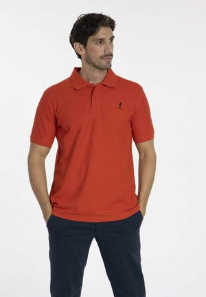 T-shirt polo rouge en tissu lisse, avec un col et des manches courtes. Petit logo noir sur la poitrine. Porté avec un pantalon foncé.