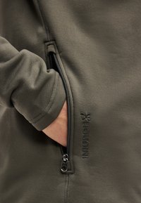 Χέρι που έχει εισαχθεί στην τσέπη με φερμουάρ του φλις μπουφάν Houdini ελιάς, εμφανίζοντας την υφή του υφάσματος και το κέντημα του λογότυπου.