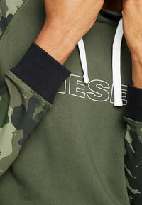 Bluza z kapturem w kolorze oliwkowej zieleni z czarnymi mankietami, wyposażona w białe sznurki i wyraźne logo "DIESEL". Rękawy w kamuflażowy wzór dodają faktury.