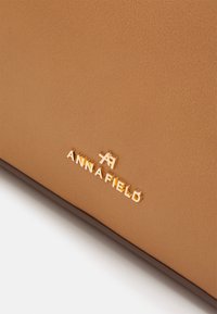 Sac à main en similicuir marron avec une texture lisse et un logo "ANNA FIELD" doré. Design simple et rectangulaire avec des accents minimaux.