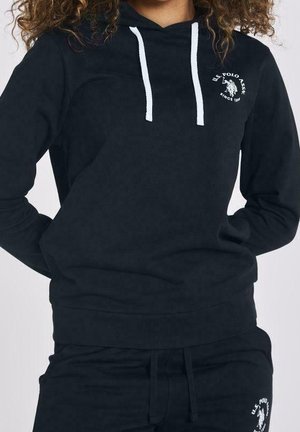 Osoba nosící černou mikinu U.S. Polo Assn. a odpovídající tepláky s bílými šňůrkami a logem na hrudi a stehně.