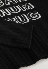 Maglione nero lavorato a maglia con testo bianco in carattere bold. La texture a coste è evidente vicino all'orlo e alle maniche.