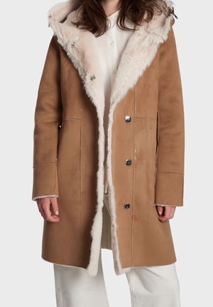 Femme portant un manteau en suède beige avec une doublure en fourrure blanche et des boutons argentés, sur une chemise blanche et un pantalon blanc, debout devant un fond uni.