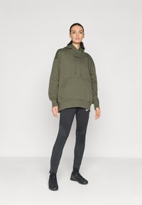 Sudadera verde oliva con bolsillo frontal y cordones, combinada con leggings negros texturizados que presentan un patrón y un logo, y zapatillas deportivas negras.