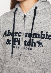 Grå zip-up hættetrøje med sorte snore og broderet "Abercrombie & Fitch New York" logo med vindtaxemblem på brystet.