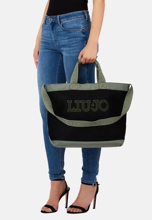 Donna con jeans blu e tacchi neri tiene una borsa a mano nera e verde oliva con il logo "LIU JO" sul davanti.