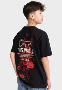En gutt med kort mørkt hår som har på seg en svart T-skjorte med rødt blomsterdesign og hvit tekst "Out of this World" på baksiden.