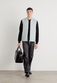 Colete acolchoado azul claro por cima de um suéter preto, combinado com jeans escuros. O modelo carrega uma bolsa de couro preta e usa sapatos pretos.