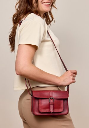 Woman wearing beige top and pants holds a small red leather crossbody bag with buckle detail at her side.