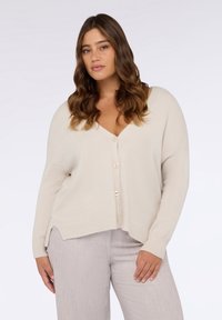 Beige Strickjacke mit V-Ausschnitt, überschnittenen Ärmeln und fünf Knöpfen vorne. Mit gerippten Bündchen und Saum sowie seitlichen Schlitzen für mehr Bewegungsfreiheit.