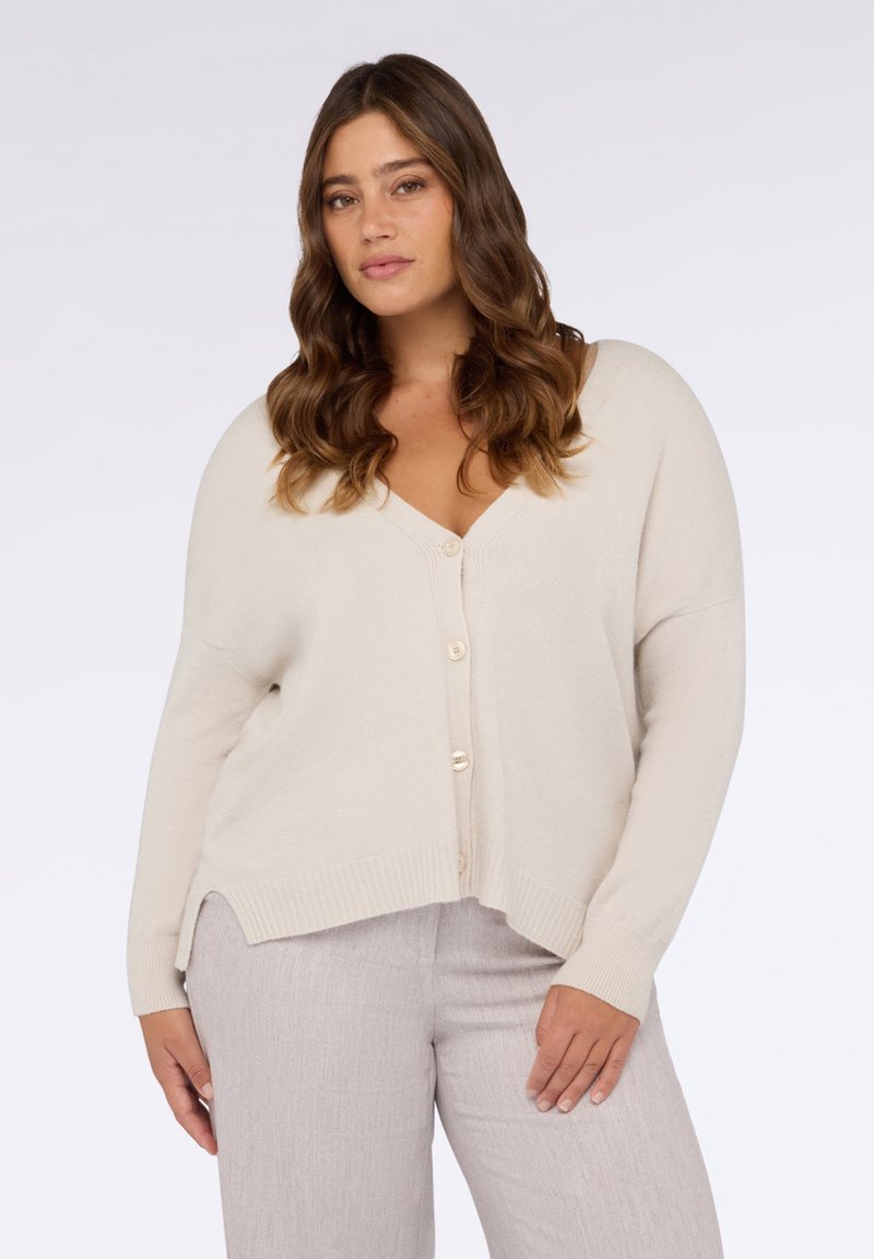 Beige Strickjacke mit V-Ausschnitt, überschnittenen Ärmeln und fünf Knöpfen vorne. Mit gerippten Bündchen und Saum sowie seitlichen Schlitzen für mehr Bewegungsfreiheit.