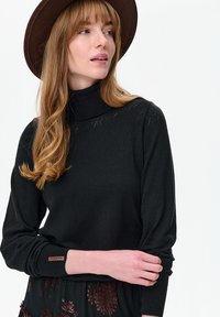 Vrouw met lang lichtbruin haar, een bruine hoed, een zwarte coltrui met patroon en een zwarte rok met bruine bloemmotieven.