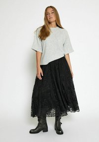 Maglione grigio a maniche corte abbinato a una gonna lunga in pizzo nera con intricati motivi floreali e stivaletti neri lucidi con tacco spesso.