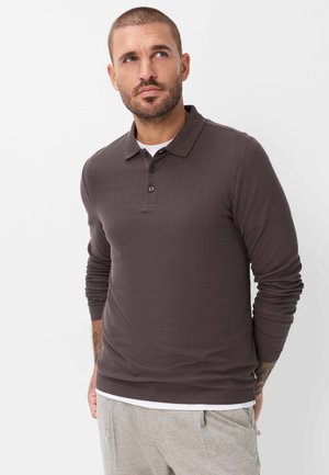 Mann mit kurzen Haaren und Bart, der ein dunkelbraunes langärmeliges Poloshirt und graue Hose trägt und seitlich vor weißem Hintergrund schaut.