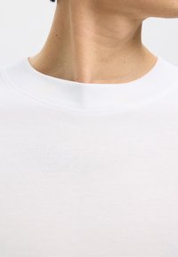 Close-up van de nek en het onderste deel van het gezicht van een persoon die een wit T-shirt met ronde hals draagt tegen een effen lichte achtergrond.