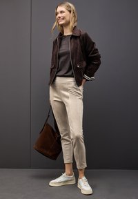 Brun mockaskinnjacka med dragkedja fram, beige tygbyxor och grå sneakers. Brun tote-väska hänger från armen. Enkel, modern design.